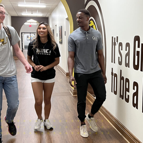 3 Quincy University students walking in a hallway with a mural that has text that sais: It is a great day to be a Hawk on it.