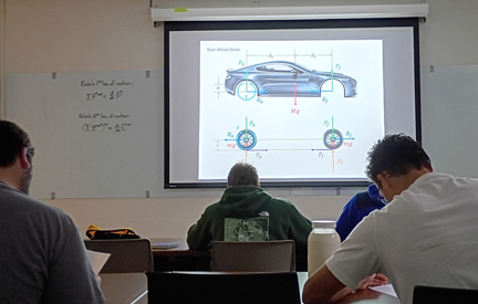 view from back of a classroom at a projection on the screen of a car and wheels with students in the foreground for an engineering class