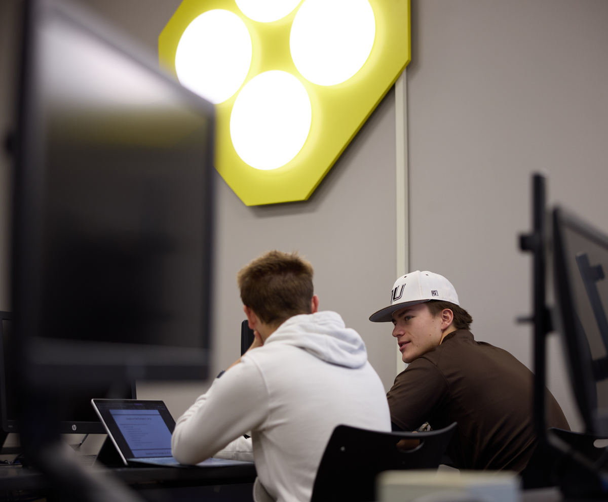 two students talking at there desk in a computer science lab