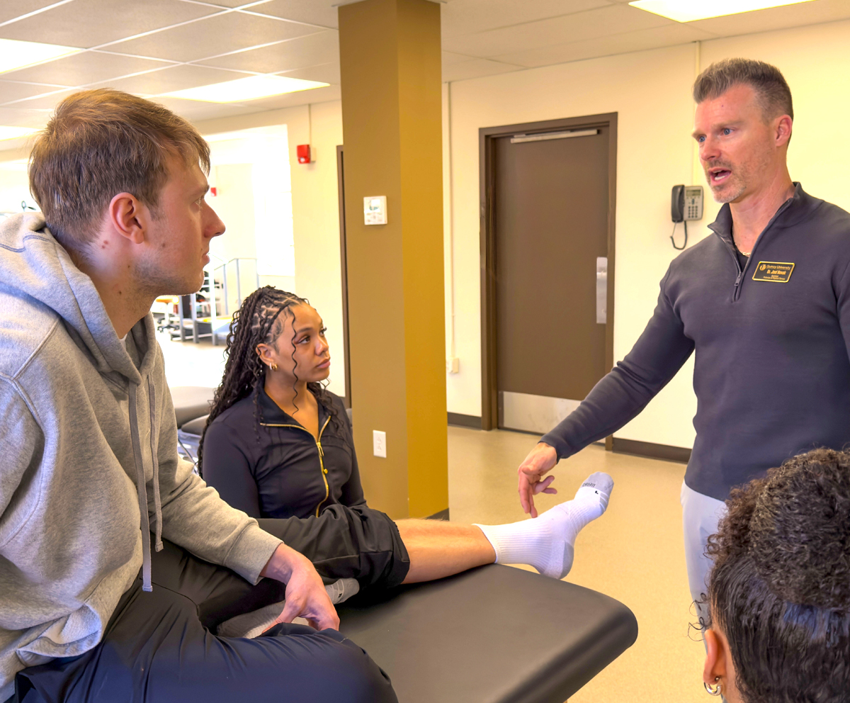 Physical therapist talking to a student who is sitting on a table and point to his foot while another student watches