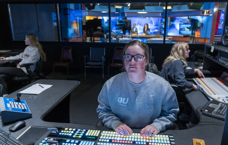student in a control room operating a switcher during a news cast. The news anchors are visible behind her.