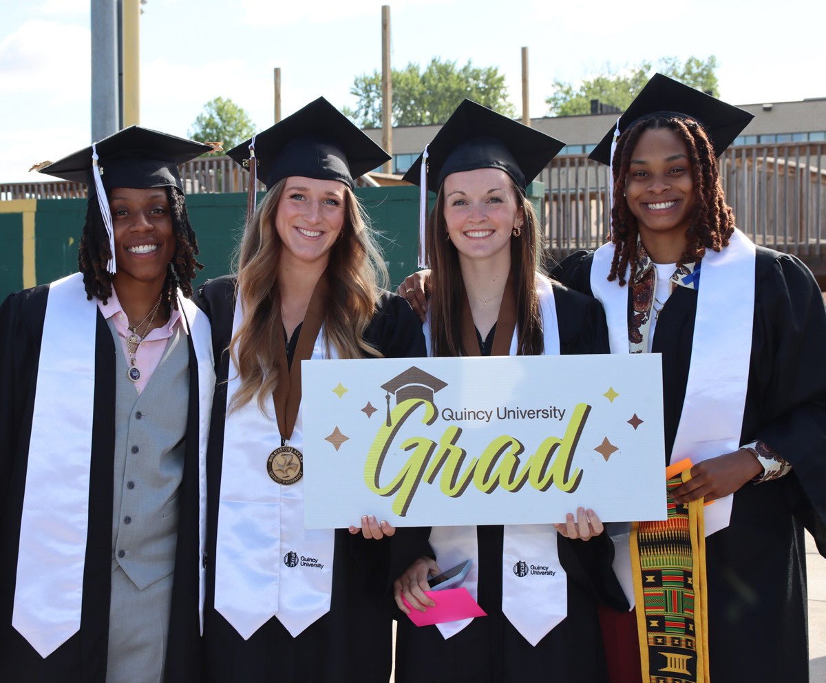 four women graduates holding a Quincy University Grad sign before graduation